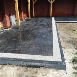 Ciemna kostka brukowa ułożona pod zadaszeniem, obramowana jasnym kamieniem, widoczne elementy konstrukcji z drewna i cegły.