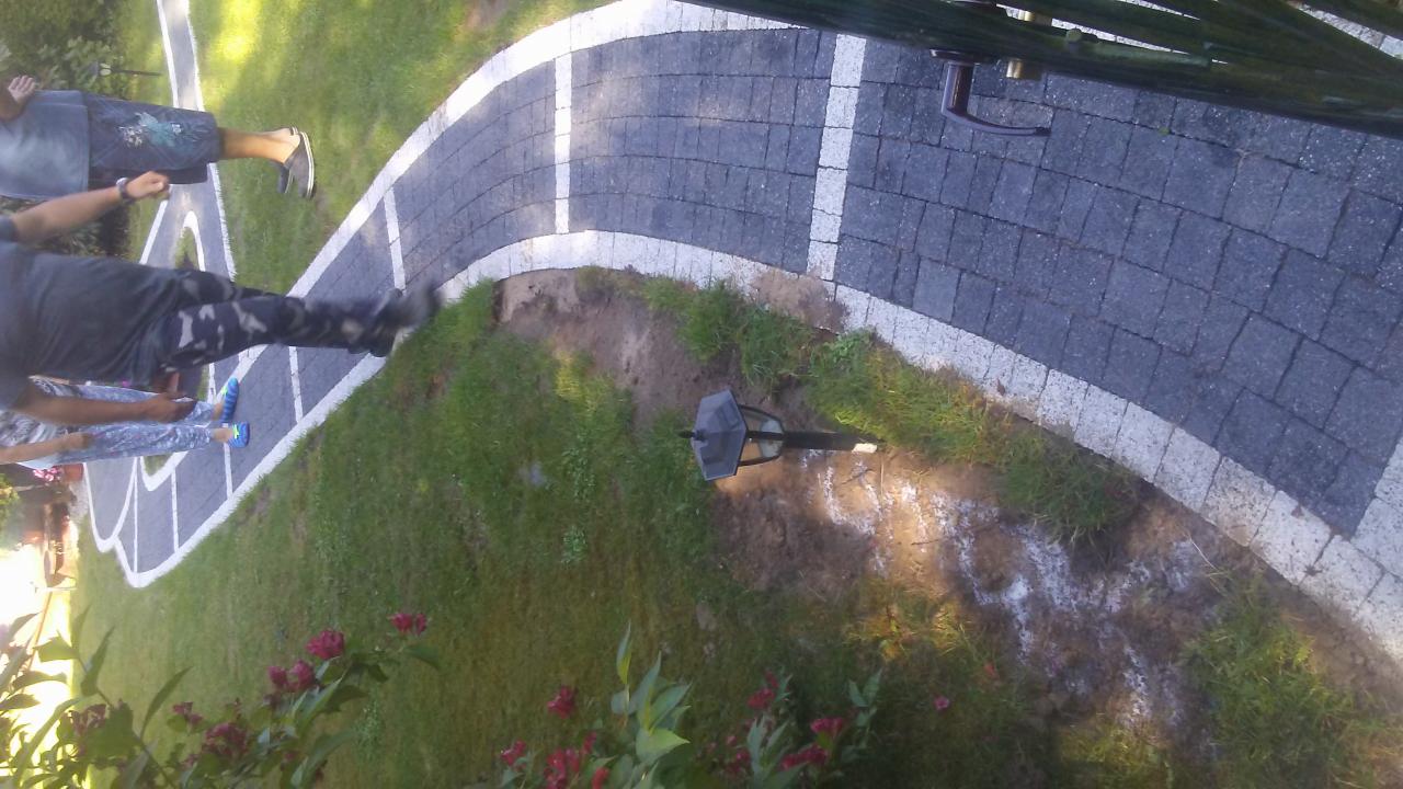 Zakrzywiona ścieżka z ciemnej kostki brukowej z białymi obrzeżami, widok z góry, w tle trawnik i osoby.