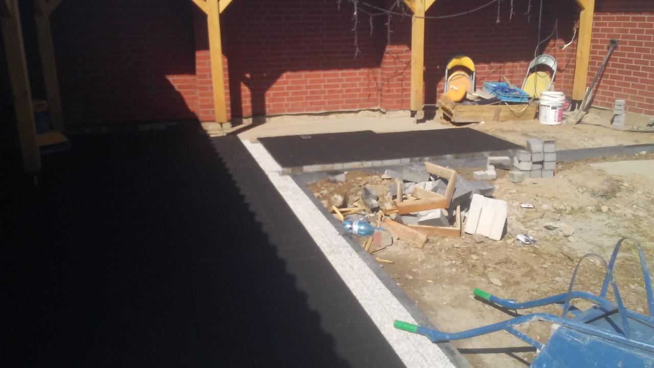 Układanie czarnej kostki brukowej z jasnoszarą obwódką, widoczne narzędzia, gruz i elementy konstrukcyjne w tle.