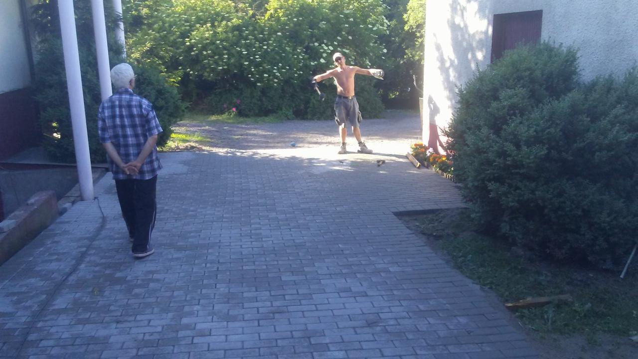 Ułożona kostka brukowa na podwórku, mężczyzna bez koszulki z rozłożonymi rękami w tle, starszy mężczyzna w koszuli w kratę obserwuje.