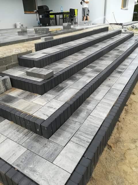 Budowa schodów zewnętrznych z kostki brukowej w odcieniach szarości, z widocznymi etapami prac i osobą w tle.