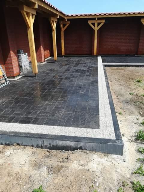 Ciemna kostka brukowa ułożona pod zadaszeniem, obramowana jasnym kamieniem, widoczne elementy konstrukcji z drewna i cegły.