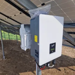 Biało-szary inwerter marki FOX podłączony do paneli słonecznych, z otwartą obudową i plastikową torbą na górze, zamontowany na metalowym słupku na ziemi.