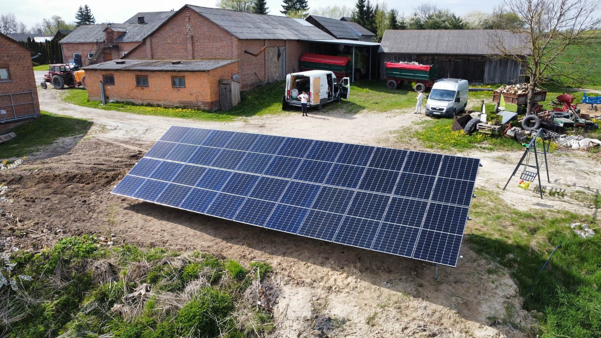 Instalacja paneli słonecznych na gruncie w wiejskim otoczeniu, widoczny samochód dostawczy i pracownicy w kombinezonach ochronnych.