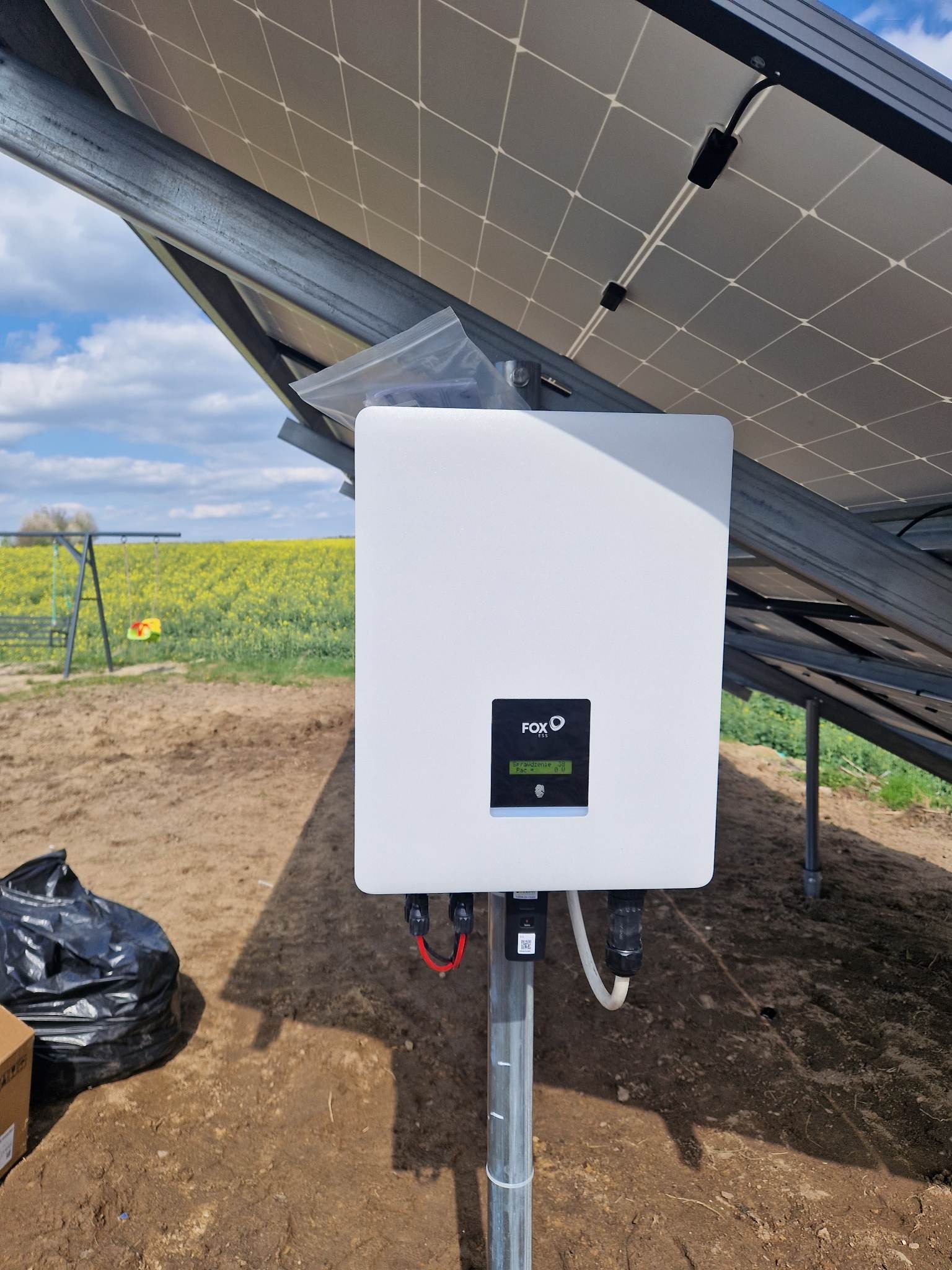 Biały inwerter FOX ESS zamontowany na metalowym słupku, pod panelami, na tle kwitnącego pola rzepaku i huśtawki.