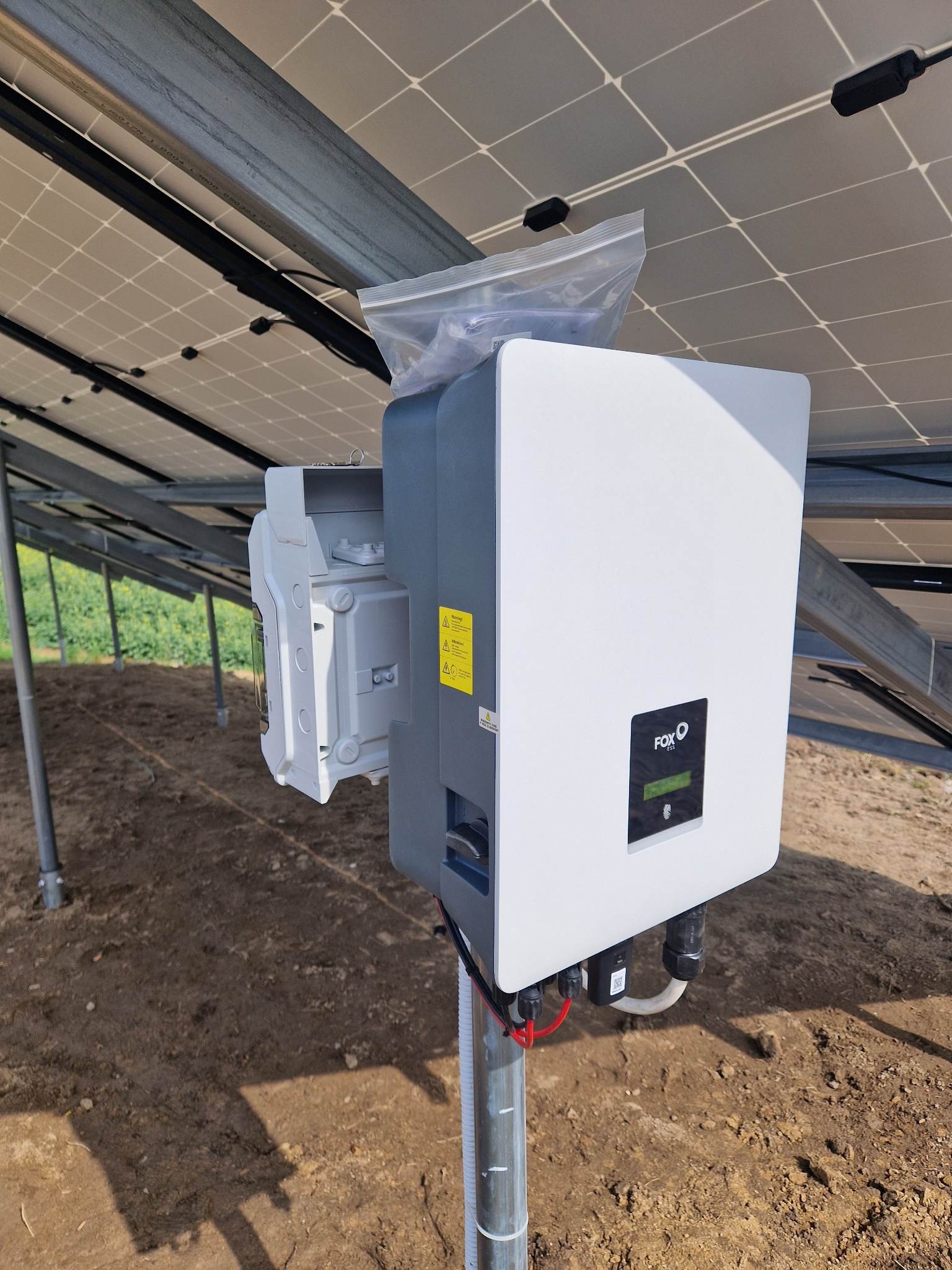 Biało-szary inwerter marki FOX podłączony do paneli słonecznych, z otwartą obudową i plastikową torbą na górze, zamontowany na metalowym słupku na ziemi.