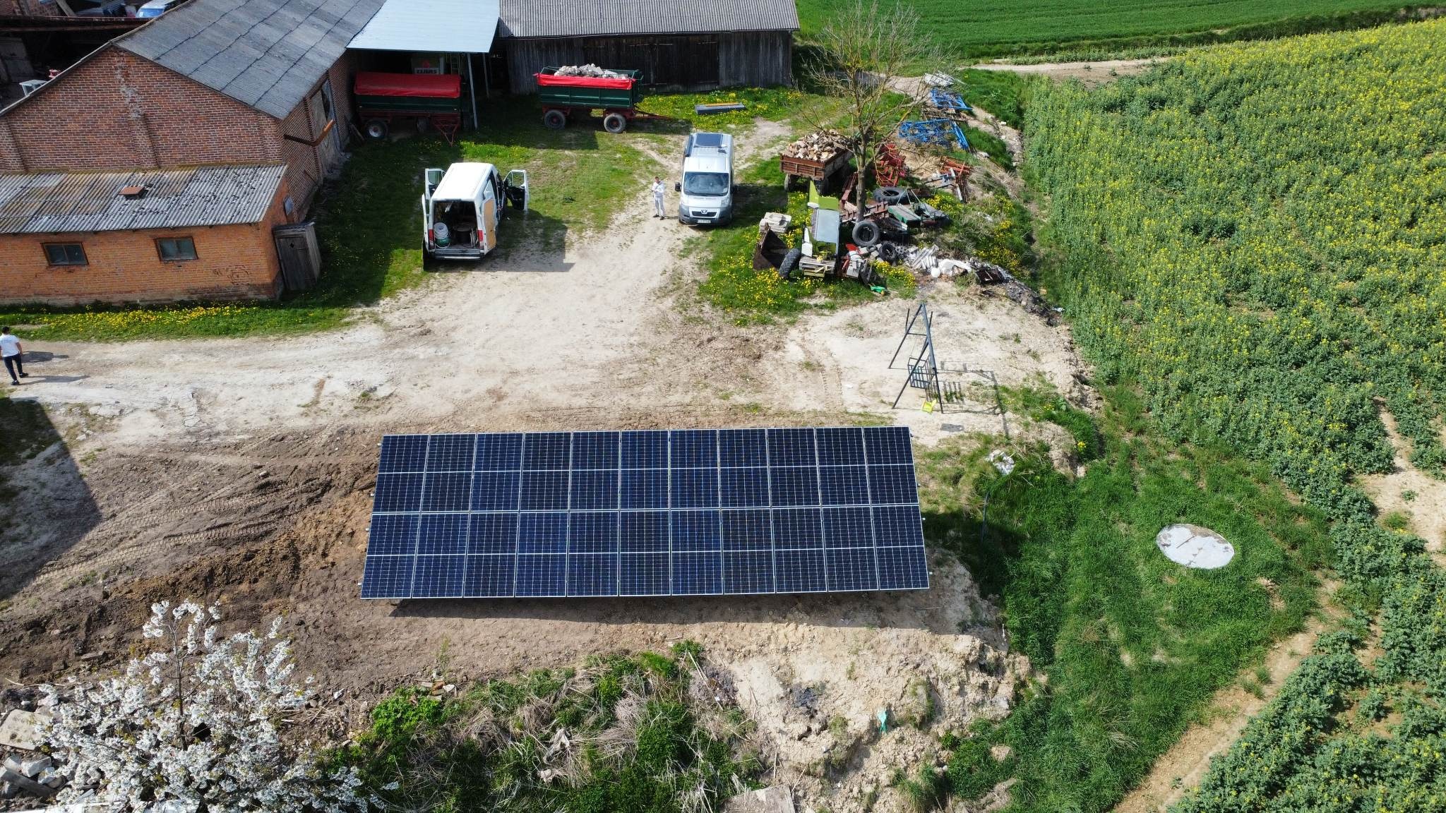 Widok z góry na instalację paneli słonecznych na gruncie w wiejskim otoczeniu, z dwoma samochodami dostawczymi i budynkami gospodarczymi w tle.