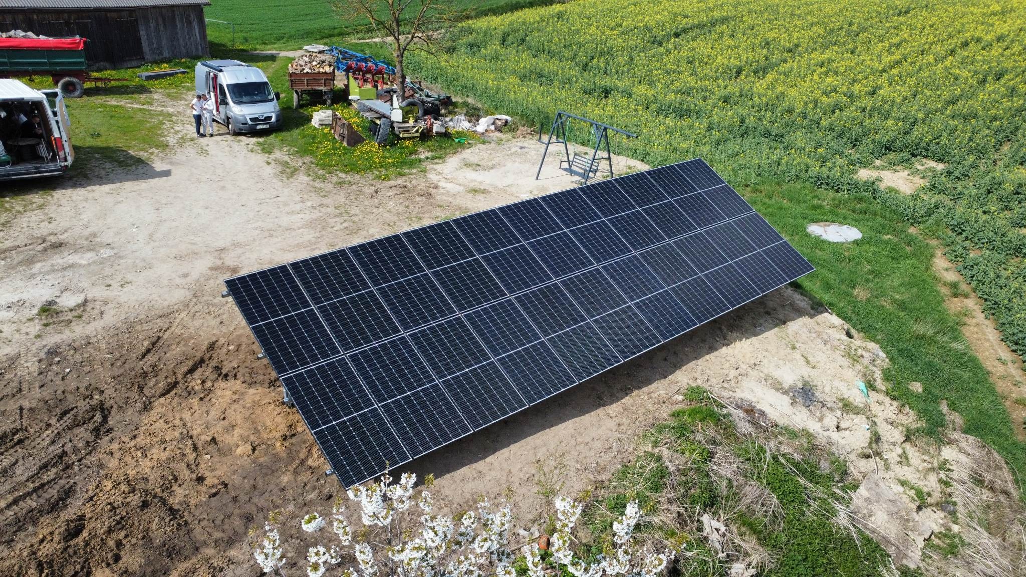 Widok z góry na nowo zainstalowane panele słoneczne na gruncie, z widocznymi dwoma samochodami dostawczymi, przyczepą z drewnem, polem rzepaku w tle oraz huśtawką.