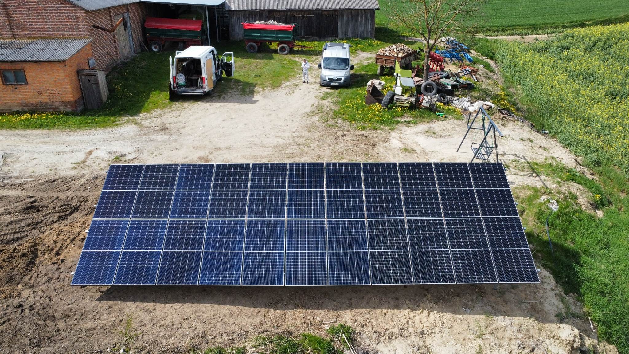 Rozległa instalacja fotowoltaiczna na ziemi, widoczna z góry, z dwoma zaparkowanymi busami i osobą w kombinezonie ochronnym w tle, obok budynków gospodarczych i pola rzepaku.