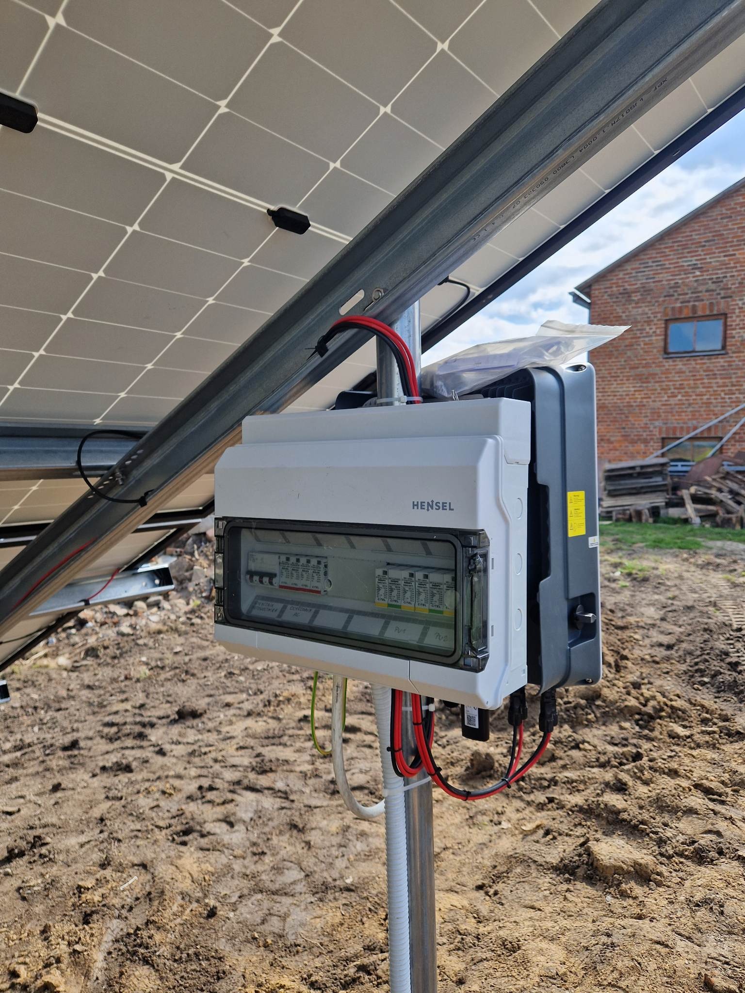 Panel słoneczny z białą skrzynką Hensel i falownikiem na metalowej konstrukcji, widoczne kable i ziemiste podłoże.