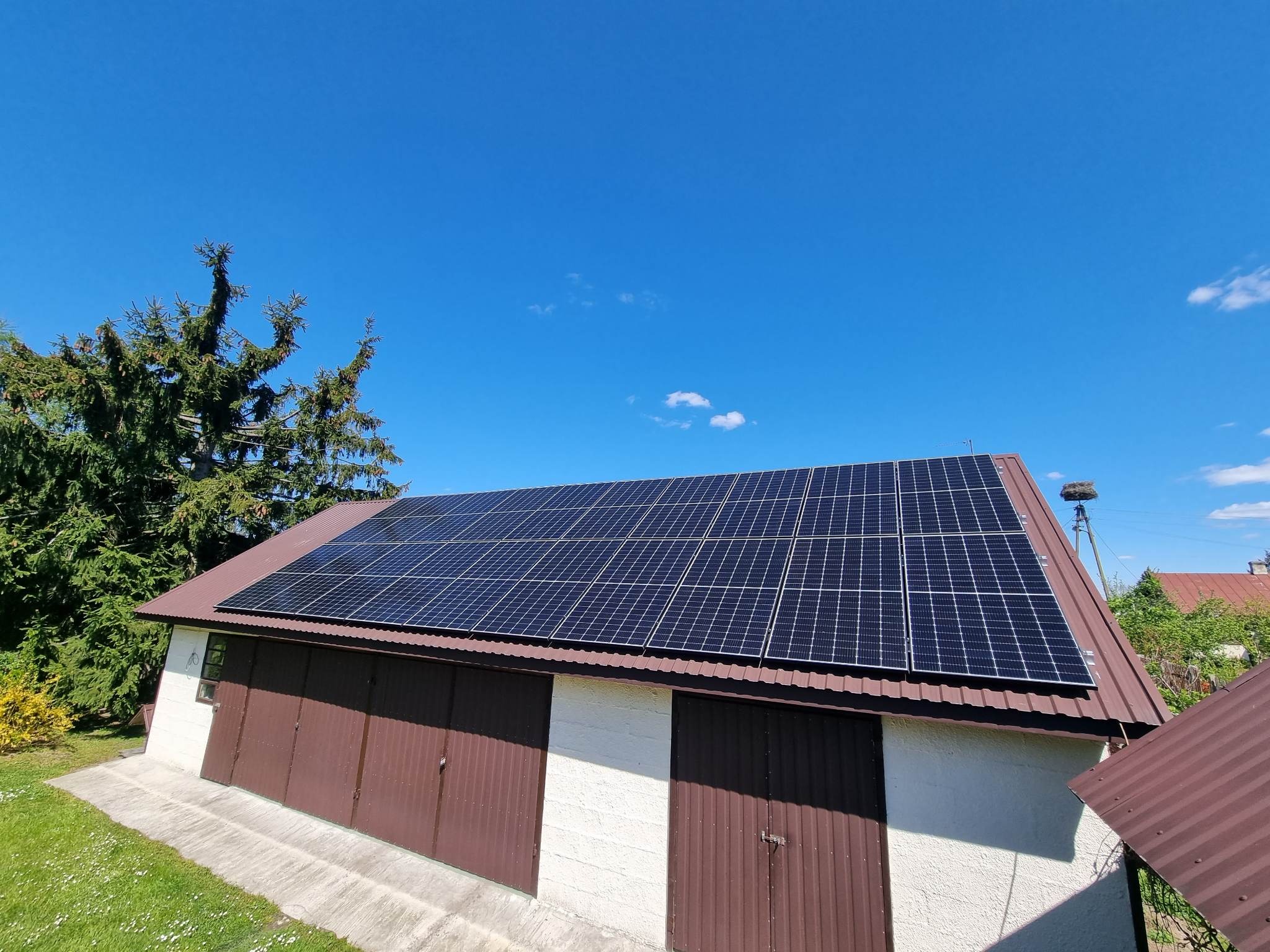 Instalacja paneli słonecznych na dachu garażu z brązowymi bramami, widok z dołu, błękitne niebo i drzewo iglaste w tle, gniazdo bocianie na słupie w oddali.