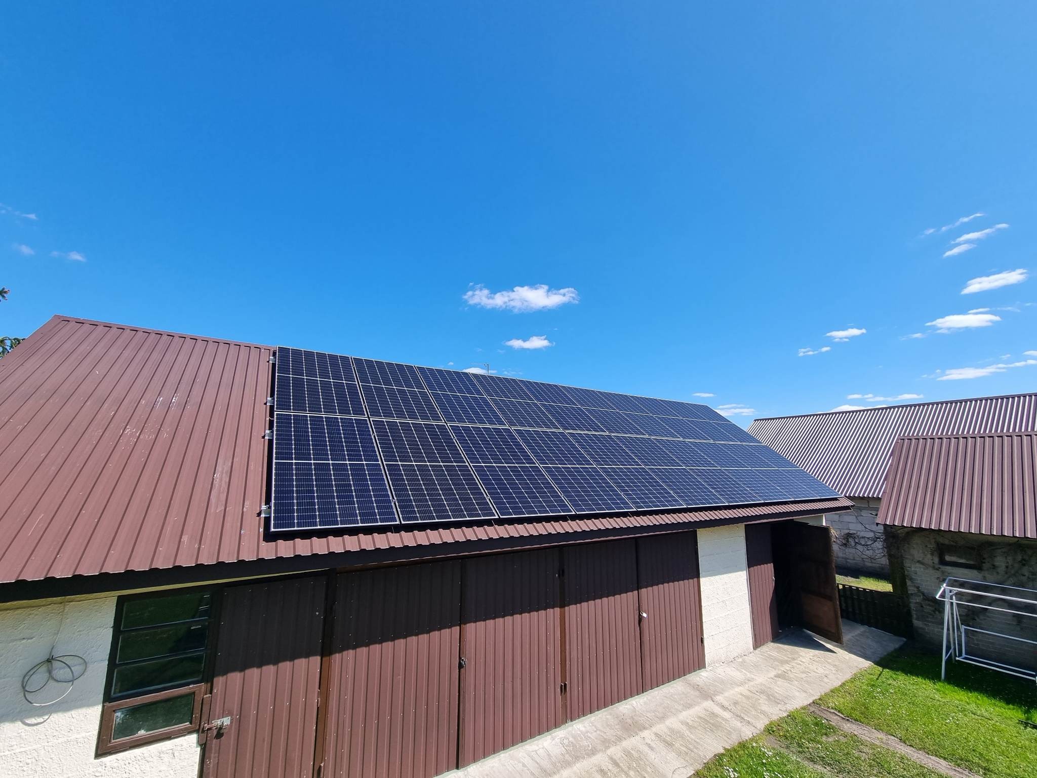 Panele słoneczne na brązowym, blaszanym dachu garażu w słoneczny dzień z błękitnym niebem i pojedynczymi chmurami.