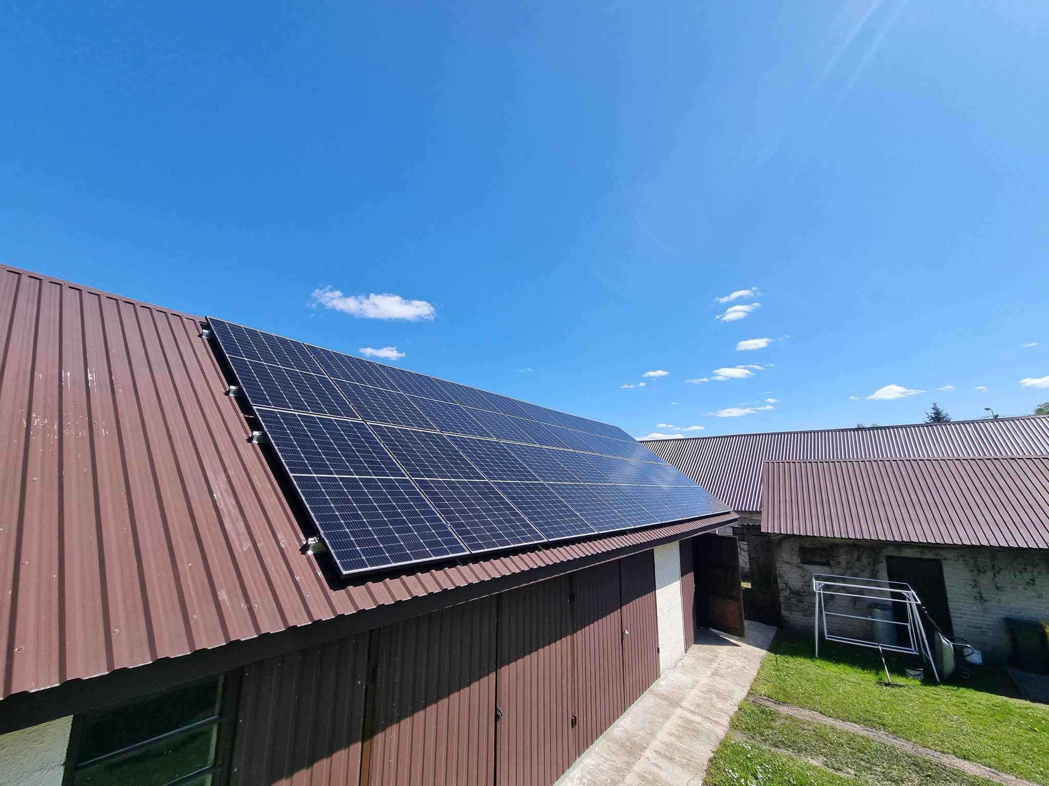 Panele słoneczne na brązowym dachu budynku gospodarczego z blachy trapezowej, błękitne niebo w tle.
