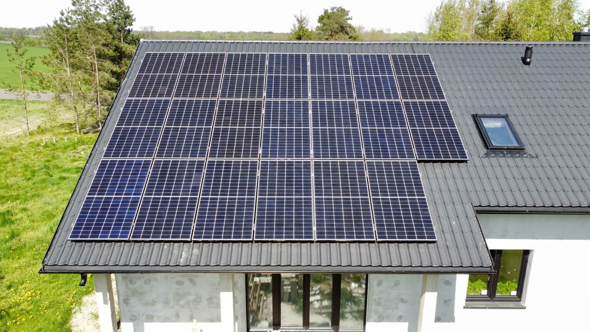 Panele słoneczne na grafitowym dachu domu jednorodzinnego z oknem dachowym, widok z góry na tle zieleni.