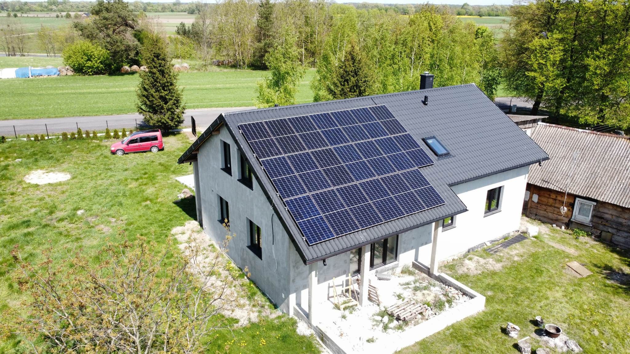 Dom jednorodzinny z szarym dachem, na którym zamontowano panele słoneczne. Z boku domu stoi czerwony samochód osobowy.