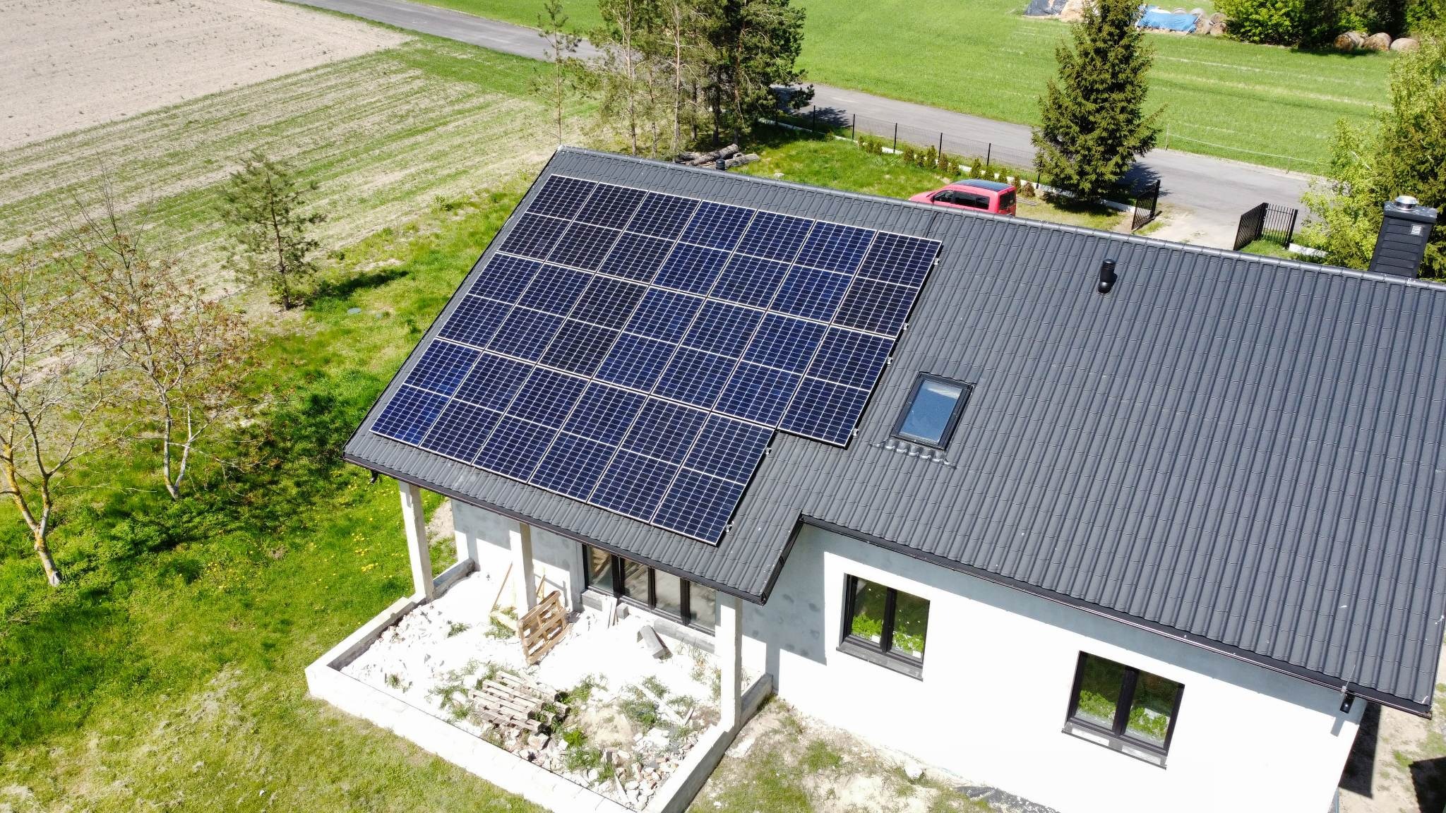 Widok z góry na dom z panelami słonecznymi na grafitowym dachu, obok czerwony samochód, na trawniku paleta budowlana, w tle pole uprawne.