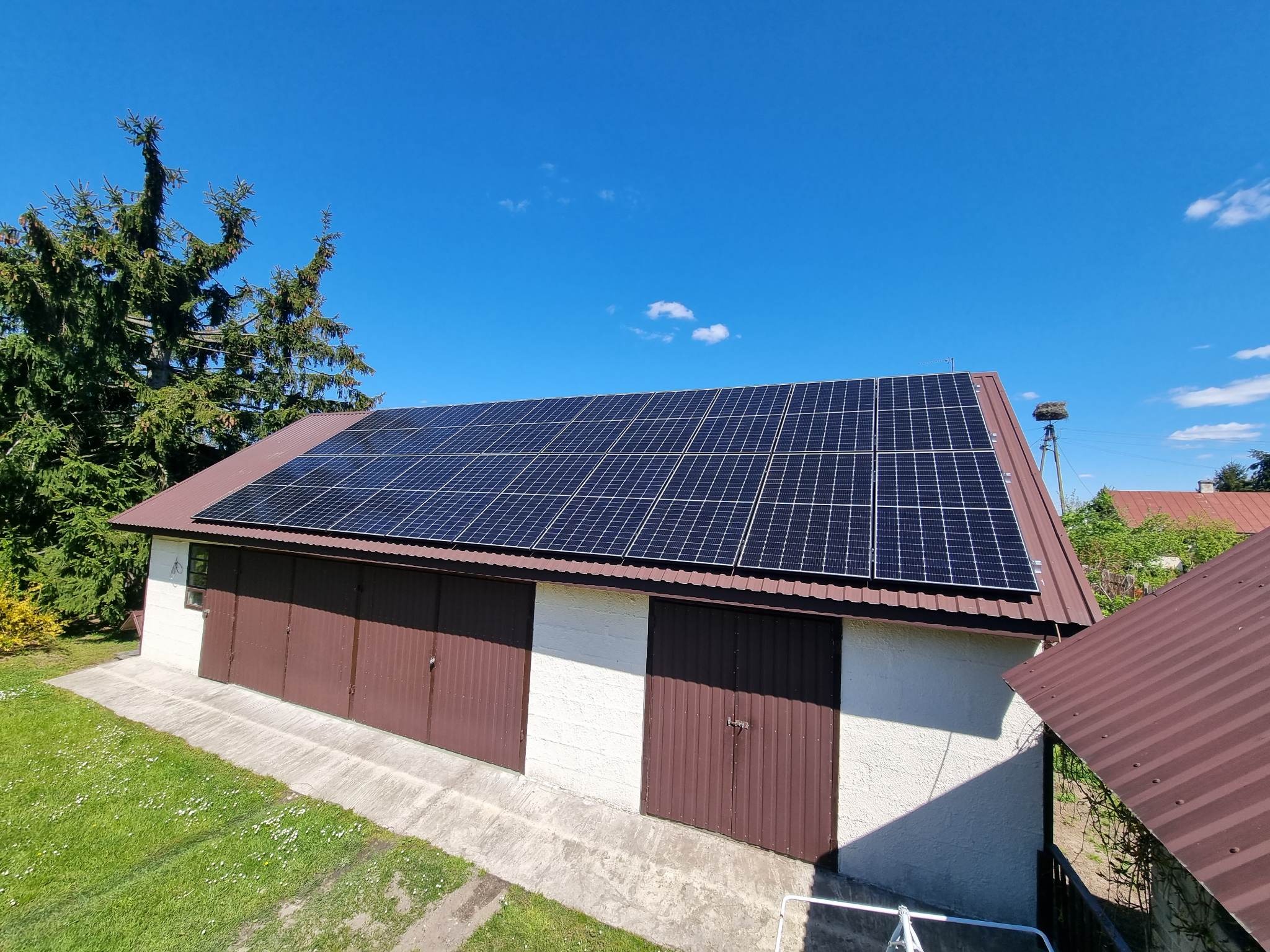 Panele słoneczne na dachu garażu z trzema brązowymi bramami, widok z góry, w tle gniazdo bocianie na słupie.