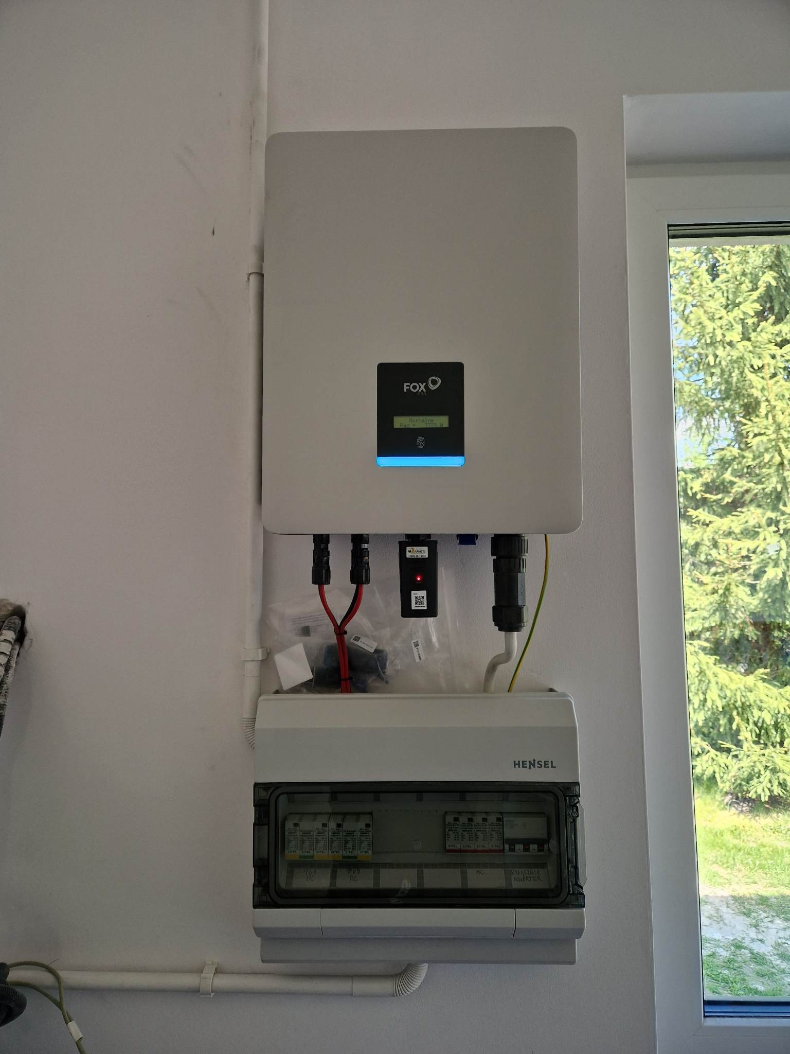 Biały inwerter marki Fox podłączony do skrzynki bezpiecznikowej Hensel na białej ścianie wewnątrz budynku, widoczne okablowanie i elementy montażowe, na wyświetlaczu inwertera widoczny napis...