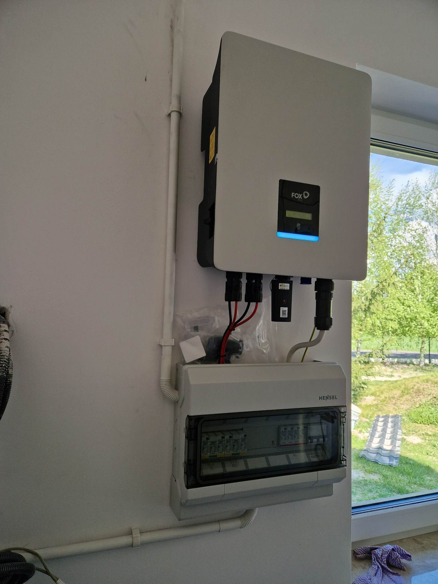 Zamontowany inwerter FoxESS obok skrzynki elektrycznej Hensel na białej ścianie, widok na zewnątrz przez okno.