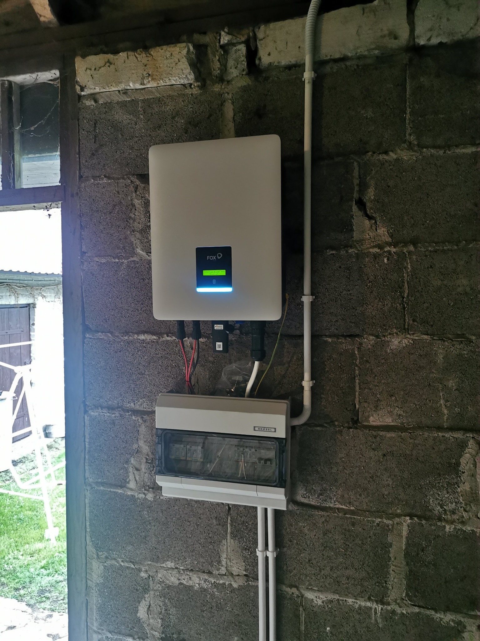Biały falownik FoxESS zamontowany na szarej ścianie z bloczków betonowych wewnątrz budynku, z widoczną skrzynką bezpiecznikową Henel i przewodami.