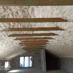 Poddasze z widocznymi drewnianymi belkami stropowymi, pokryte grubą warstwą białej pianki izolacyjnej, ściany z szarego bloczka betonowego, okno z folią zabezpieczającą.