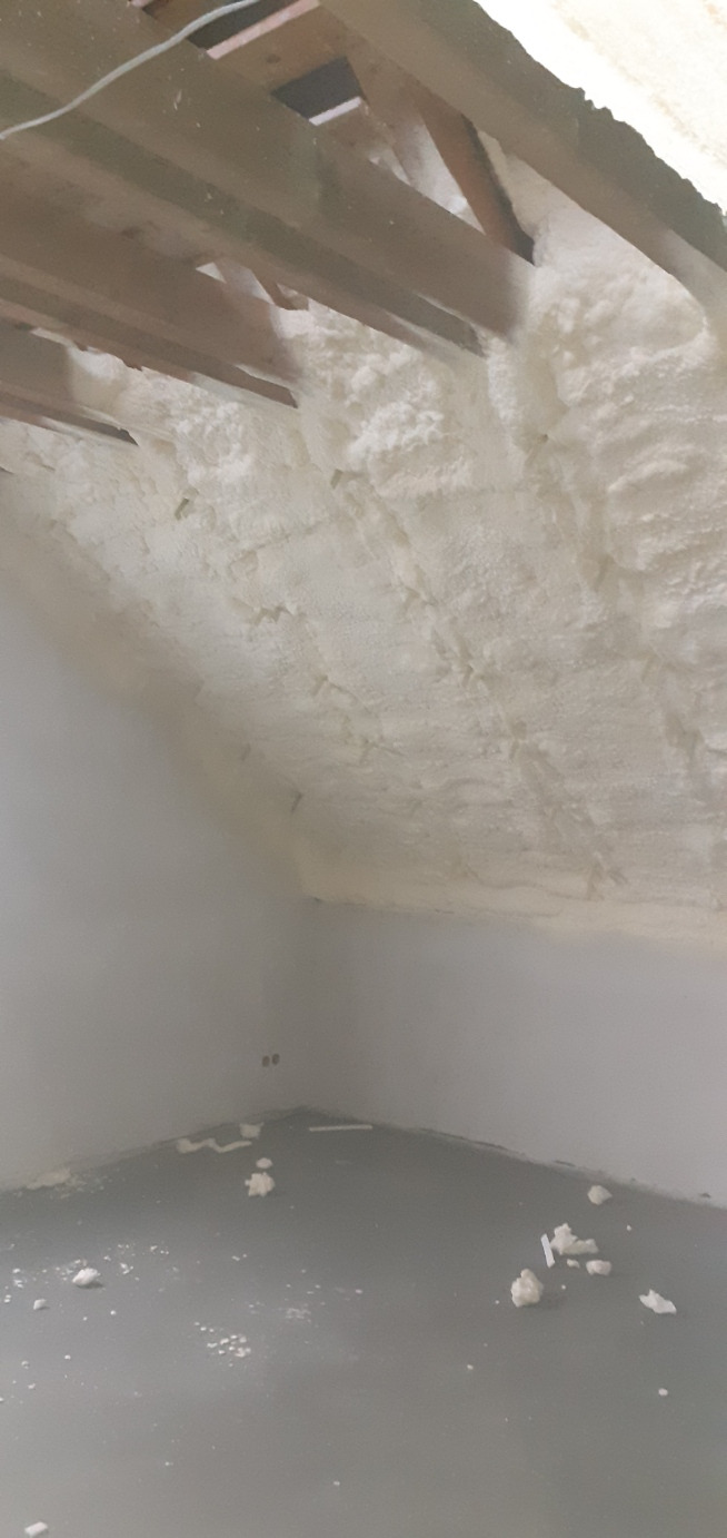 Poddasze z widocznymi krokwiami, pokryte grubą warstwą białej pianki izolacyjnej, z resztkami pianki na szarej podłodze.