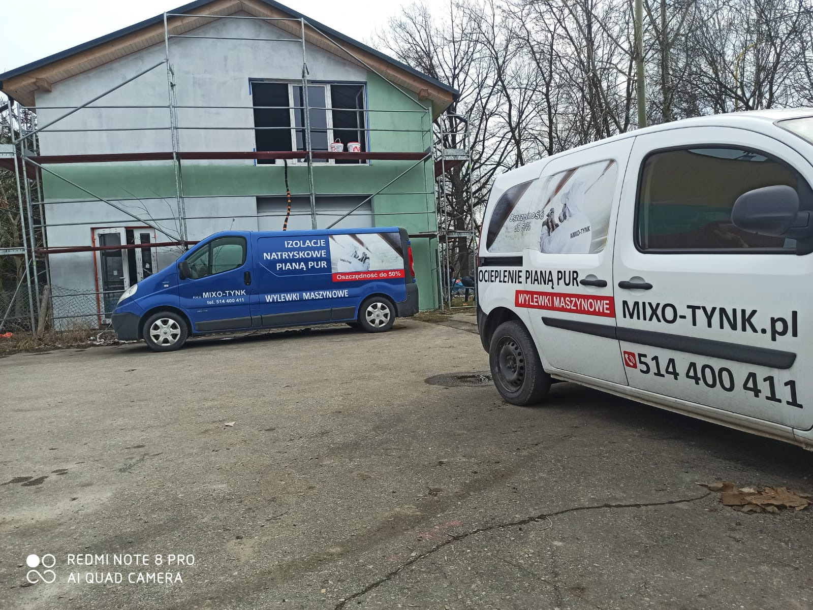 Dwa busy firmy Mixo-Tynk z reklamą izolacji natryskowej pianą PUR i wylewek maszynowych zaparkowane przed ocieplanym domem ze stojącym rusztowaniem.