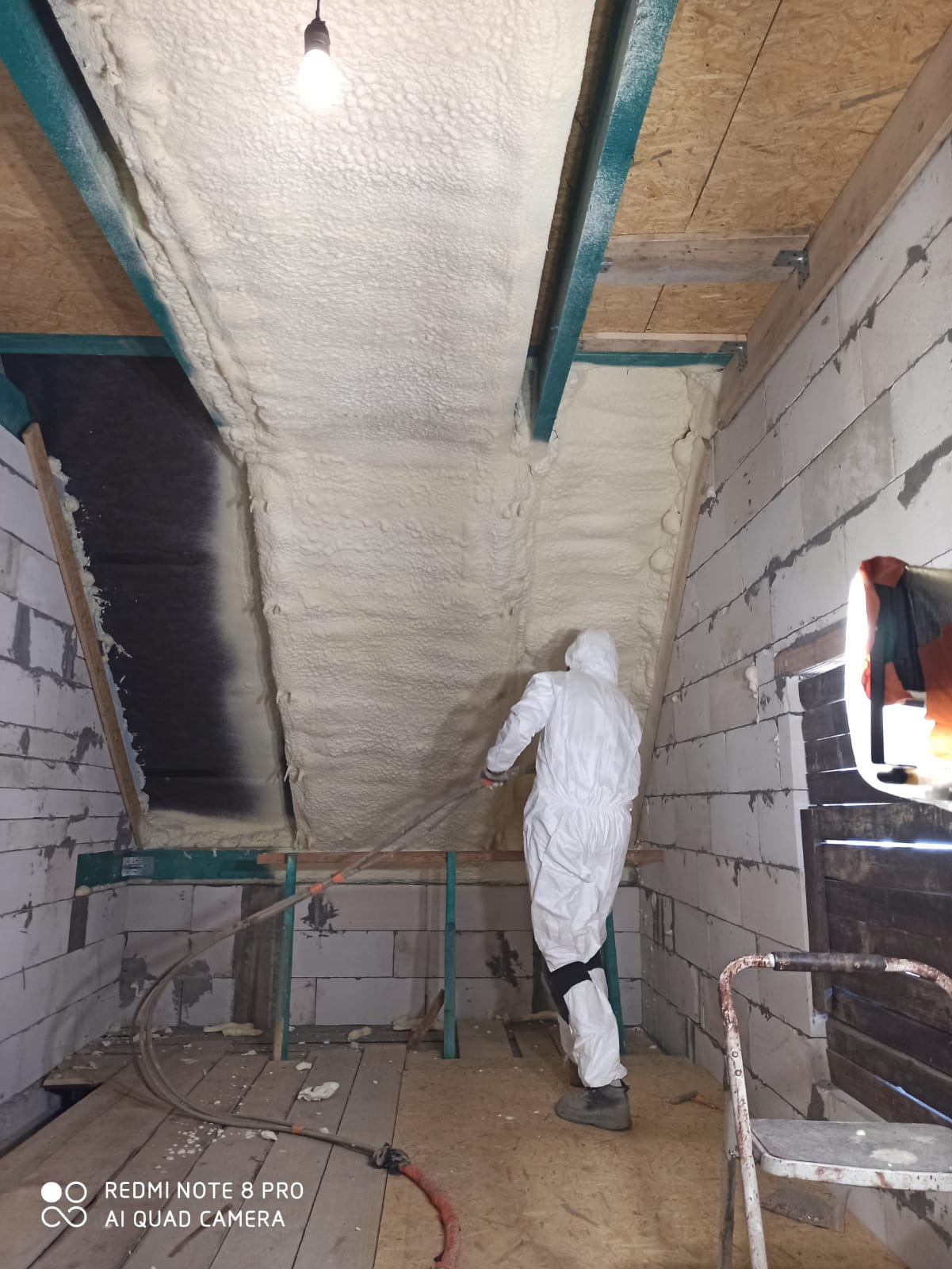 Pracownik w kombinezonie ochronnym natryskuje pianę izolacyjną na poddasze z widocznymi elementami konstrukcyjnymi, takimi jak belki pomalowane na zielono i białe ściany z pustaków.