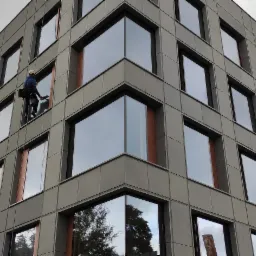 Dwóch alpinistów przemysłowych myje okna na elewacji nowoczesnego biurowca z szarymi panelami i dużymi, lustrzanymi szybami.