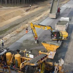 Żółty kombajn drogowy i koparka JCB w trakcie budowy drogi, z ciężarówkami dowożącymi materiał i pracownikami w odblaskowych ubraniach nadzorującymi proces.