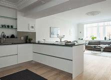 Jasne, minimalistyczne wnętrze: biała kuchnia z czarnym blatem przechodząca w salon z szarą sofą, drewniana podłoga, duże okno z widokiem na zieleń, dekoracyjny sufit.