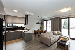 Jasne, przestronne wnętrze nowoczesnego salonu z aneksem kuchennym. Szare meble kuchenne z czarnymi akcentami, drewniany stół z krzesłami, beżowa sofa i stolik kawowy. Duże okna przesuwne...