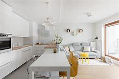 Jasne, minimalistyczne wnętrze: biała kuchnia z drewnianym blatem przechodząca w salon z szarą sofą i żółtym stolikiem. Widoczne dekoracje ścienne i duże okno balkonowe.