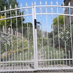 Otwarta, dwuskrzydłowa, metalowa brama z dekoracyjnymi elementami i siatką, prowadząca do ogrodu z kwitnącymi roślinami.