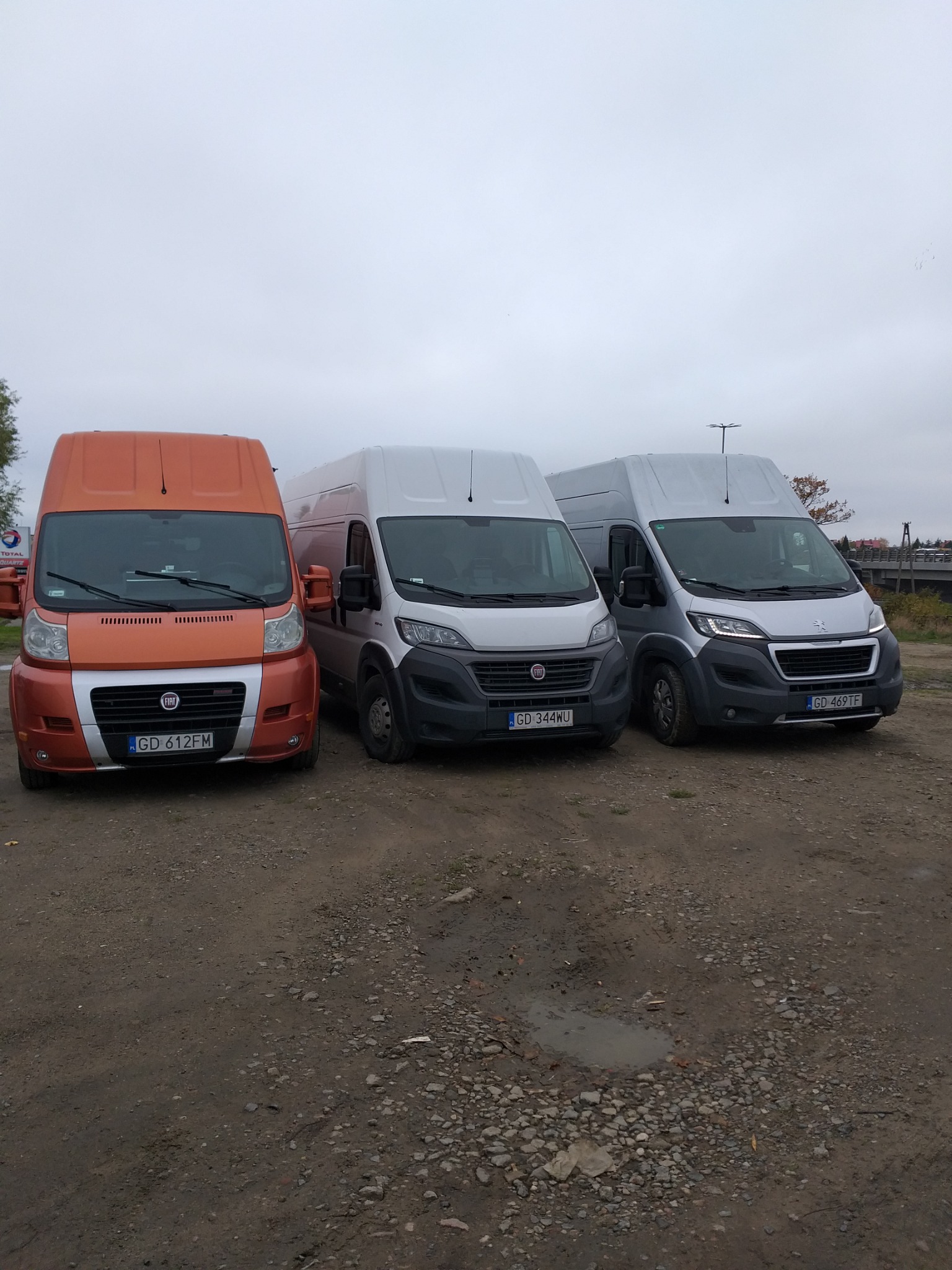 Trzy busy marki Fiat i Peugeot stojące na żwirowym placu, pomarańczowy z przodu, dwa srebrne za nim, widoczne tablice rejestracyjne.