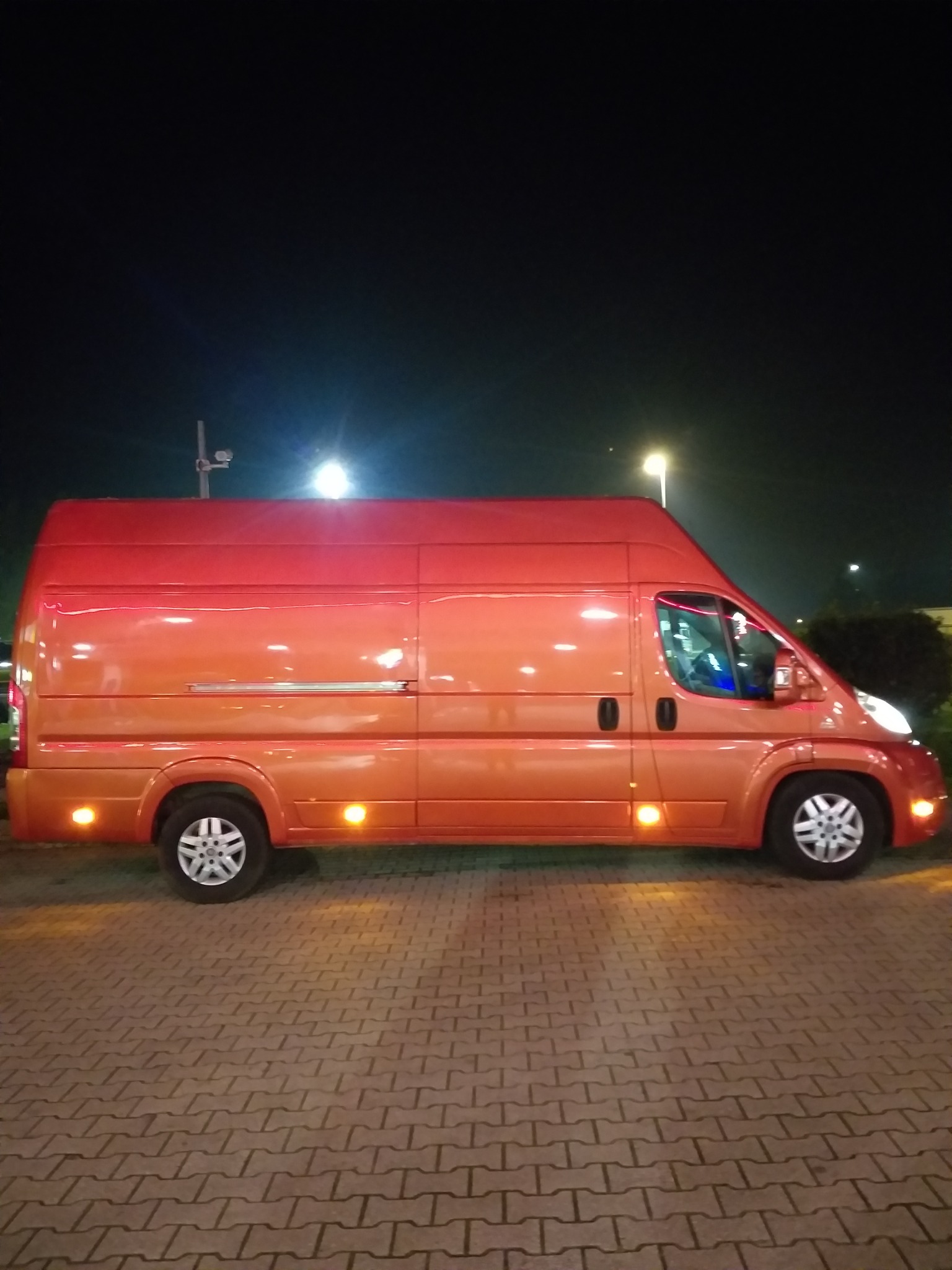 Pomarańczowy bus dostawczy zaparkowany nocą na brukowanym placu, oświetlony światłami ulicznymi.