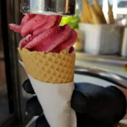 Nakładanie różowego, kręconego loda z automatu do wafelka trzymanego w rękawicy ochronnej, w tle pojemnik z wafelkami.