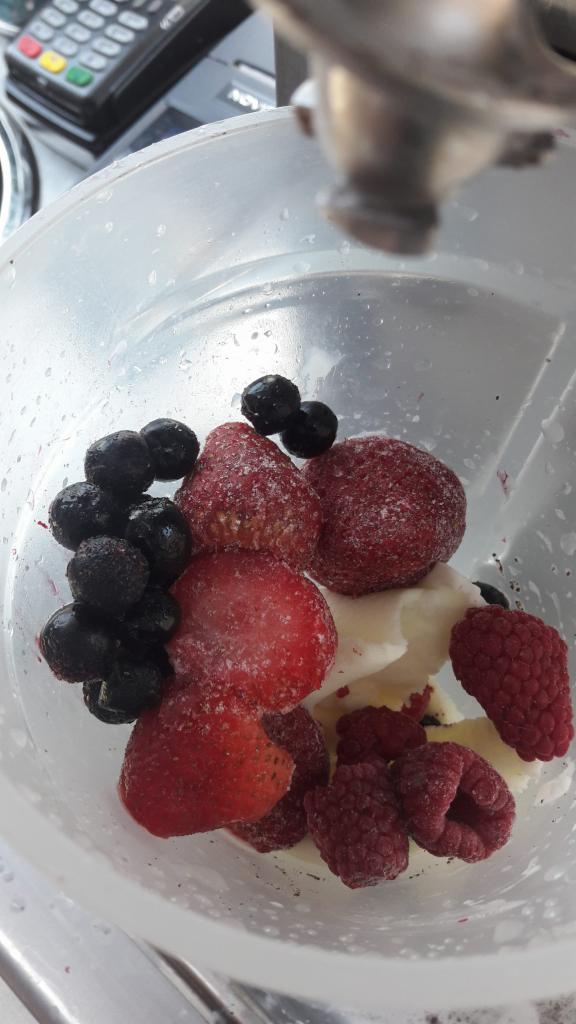 Mrożone truskawki, maliny i borówki w plastikowej misce, przygotowywane do deseru, z terminalem płatniczym w tle.