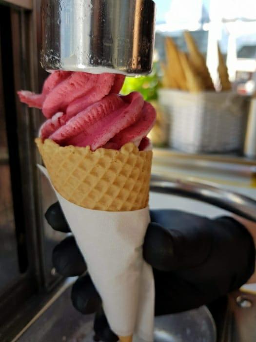 Nakładanie różowego, kręconego loda z automatu do wafelka trzymanego w rękawicy ochronnej, w tle pojemnik z wafelkami.