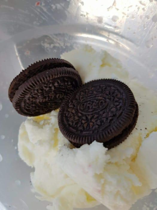 Dwa ciastka Oreo ułożone na porcji białych lodów w plastikowym pojemniku, widok z góry.