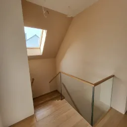 Schody drewniane z szklaną balustradą w nowo wykończonym domu, widok z góry na dół, z widocznym oknem dachowym i wystającymi kablami elektrycznymi.