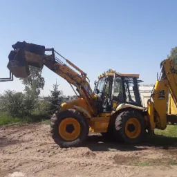 Żółta koparko-ładowarka JCB na nierównym, piaszczystym terenie z widocznymi śladami opon, pod błękitnym niebem. Z przodu łyżka z widłami, z tyłu ramię koparki.
