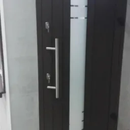 Ciemna brama garażowa z pionowym, matowym przeszkleniem, wyposażona w długi, metalowy uchwyt i dwa zamki, ustawiona w surowym, betonowym otoczeniu.