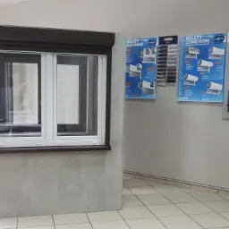 Ekspozycja okna z brązową roletą zewnętrzną nadstawną w salonie, obok plansze reklamowe rolet i mechanizmów.