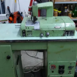 Zielona, przemysłowa maszyna do szycia skór, widoczne ślady użytkowania, ustawiona na roboczym stole w warsztacie.