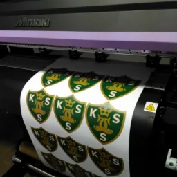 Drukarka Mimaki drukuje arkusz naklejek z zielono-złotym herbem KSS na białym tle.