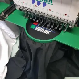 Proces haftowania logo 'PLUS REKLAM' na czarnej koszulce przy użyciu maszyny Tajima z zielonym ramieniem, widoczne nici i igły w trakcie pracy.