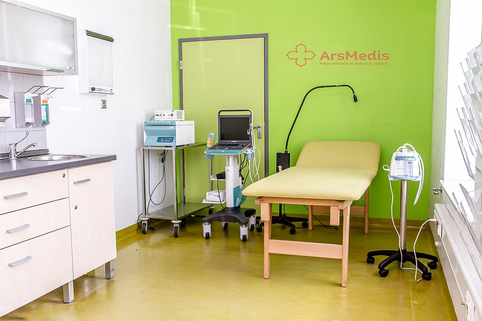 Jasne wnętrze gabinetu lekarskiego z kozetką, aparaturą medyczną i umywalką, logo ArsMedis na zielonej ścianie.