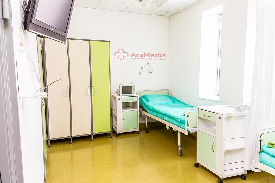Jasny pokój w klinice z łóżkiem szpitalnym, szafkami nocnymi i telewizorem na ścianie, logo ArsMedis na ścianie.