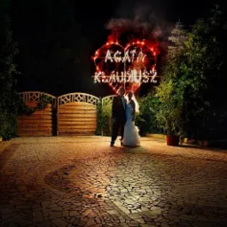 Para młoda stoi na tle płonącego serca z fajerwerków z ich imionami na tle drewnianej bramy i zieleni.